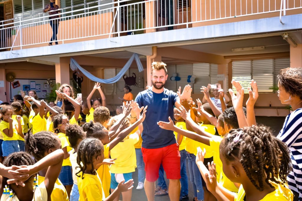 Louis Margot, souriant et entouré d'un groupe d'enfants enthousiastes vêtus de jaune, lève les bras dans une cour d'école sous le soleil radieux de la Martinique.